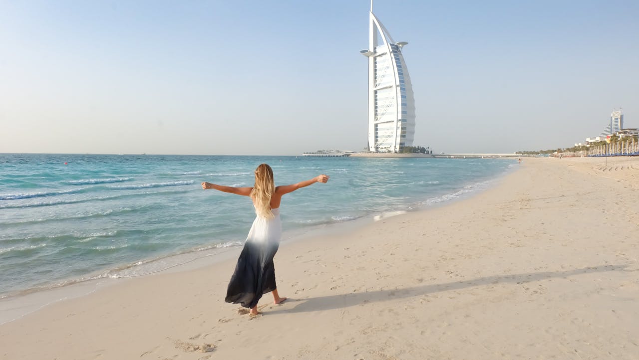 Home A carefree woman in a flowing dress strolls along the Dubai beach with Burj Al Arab in view.